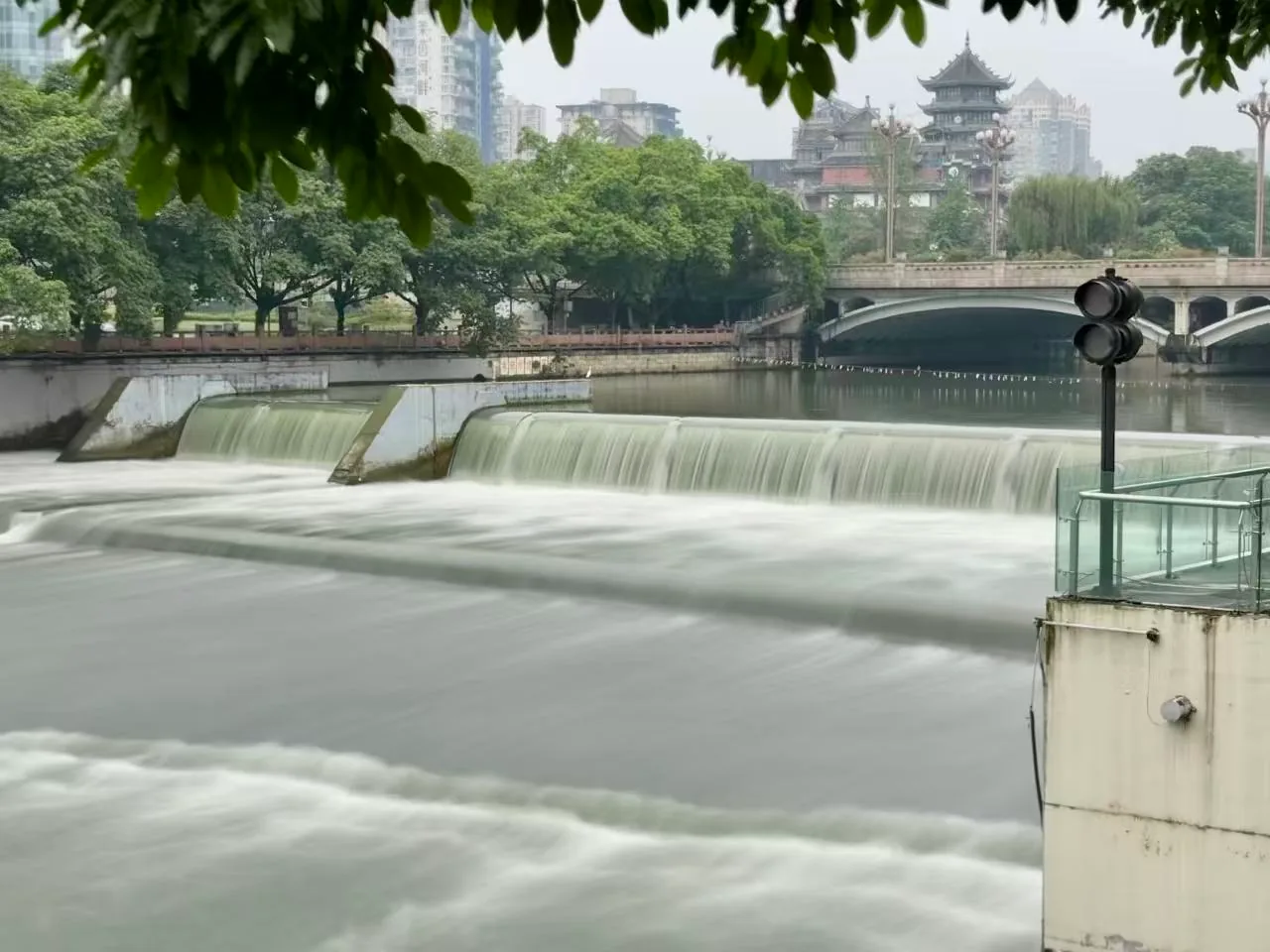 城市河道上的小水坝，远处有桥和楼。